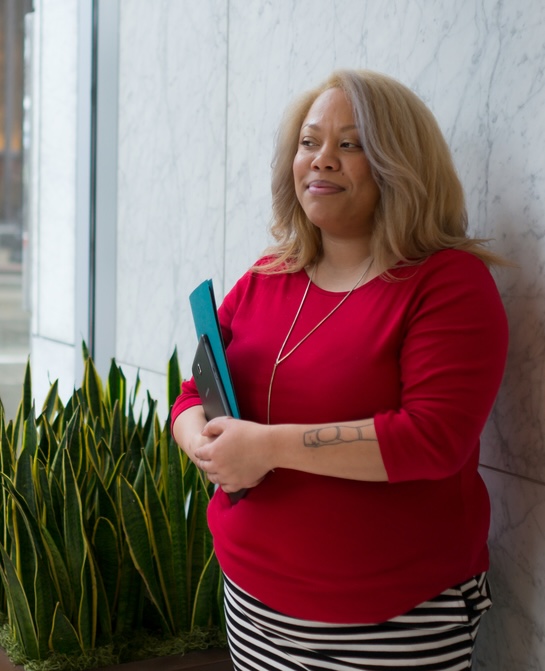 An African-American lesbian woman stands in front of a white and gray marble stone wall in an upscale office building, holding a tablet computer and a blue paper folder. She's wearing a red knit top with elbow-length sleeves, a long silver necklace, a black-and-white-striped pencil skirt and red patent high heeled shoes. Her skin is medium brown and she has blonde hair and a tattoo on one arm.
