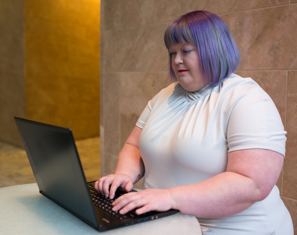 Plus-Size Businesswoman in Office Building A fat white woman sits in a large, formal office building at a high table and uses a laptop computer. She's wearing a light gray dress with tan high-heeled shoes and has a visible belly outline. Her hair is purple and she has pale skin.