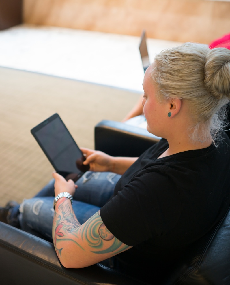 Two women work on a tablet and laptop computer as they sit in black leather-like armchairs in an upscale office building lobby. A blue folder and papers rest on a table between them. The plus size, African American lesbian woman is wearing a red knit top with elbow-length sleeves, a long silver necklace, a black-and-white-striped pencil skirt and red patent high heeled shoes. Her skin is medium brown and she has blonde hair and a tattoo on one arm. The white lesbian woman is casually dressed in a black t-shirt, ripped blue denim jeans, a silver watch and black ankle boots. She has a colorful full sleeve tattoo and pale skin. Her blonde hair is pulled up in a bun. The floor of the building is white/gray marble and the walls are dark cherry wood and white marble. The area where they're sitting has a rug on the floor as well as a black marble and metal side table and another chair. On the other side is a plant and a plate glass window looking out onto the street.